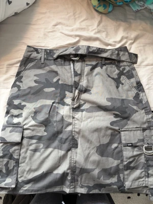 Camouflage kjol från Vans med fickor - Säljer en grå camouflagekjol från Vans med praktiska cargofickor och bälte i midjan. Kjolen har en cool streetstil och är perfekt för dig som gillar utility-vibbar. Tillverkad i bomull för skön känsla och slitstyrka.