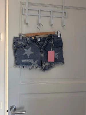 Blå jeansshorts med stjärnor - Säljer ett par blå lågmidjade jeansshorts med ljusa stjärnor som mönster. Shortsen har fransig kant, klassisk femficksmodell och dragkedja. Perfekta för sommaren och ger en cool, avslappnad vibe.
