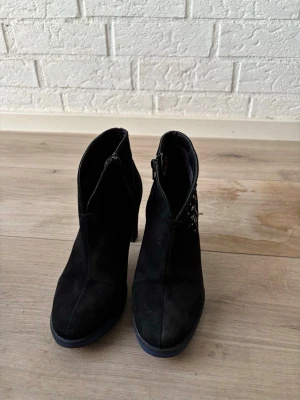 Svarta stövletter från Desigual - Svarta stövletter från Desigual med dekorativa färgade stenar på sidan och broderad logga. Skorna har en hög klack, rundad tå och dragkedja på insidan. Materialet är mocka-liknande och ger en mjuk känsla. Perfekt för dig som vill ha något unikt och snyggt.