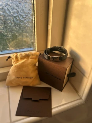 Louis Vuitton svart läderarmband - Stilrent svart armband från Louis Vuitton i läder med silverfärgat spänne och LV-logga framtill. Armbandet har justerbart spänne och kommer med originalpåse och ask. Perfekt accessoar för att lyfta din outfit.