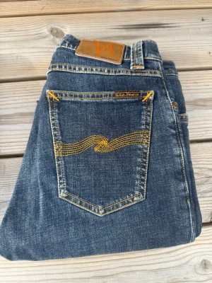 Blå jeans från Nudie Jeans - Säljer ett par klassiska blå jeans från Nudie Jeans med snygga gulbroderade detaljer på bakfickorna och läderpatch i midjan. Jeansen har rak passform och är tillverkade i slitstarkt denim-material. Perfekta för dig som gillar stilrena och tidlösa jeans.