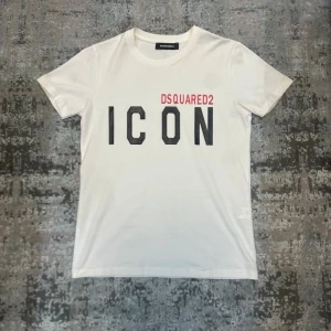 Vit Dsquared  t-shirt - Säljer en vit t-shirt med stort svart ICON-tryck text på bröstet. Klassisk rund hals och korta ärmar. Perfekt för dig som vill ha en stilren och trendig look. Aldrig använd endast testad