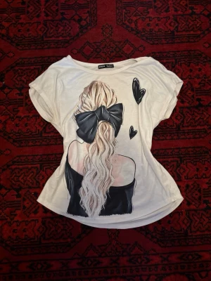 Vit t-shirt med tryck  - Vit t-shirt från Green Coast med korta ärmar och ett stort tryck på ryggen av en tjej med långt blont hår och en svart rosett. Två svarta hjärtan är också tryckta på ryggen. T-shirten har en avslappnad passform och är gjord i mjuk bomull.