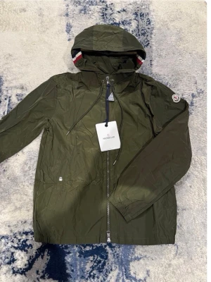 Helt ny Grön Moncler vindjacka med huva med paket och påse  - Snygg grön vindjacka från Moncler med huva och dragkedja framtill. Jackan har Moncler-logga på ärmen och coola rödvita detaljer på insidan av huvan. Materialet är lätt och vattenavvisande, perfekt för blåsiga dagar. Jackan har elastiska ärmslut och två fickor.