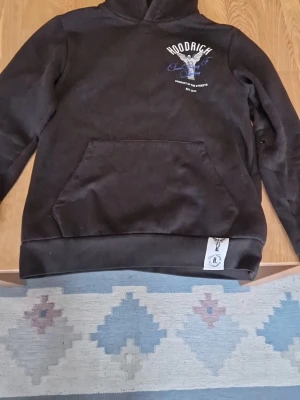 Svart hoodie från Hoodrich med ängelmotiv - Säljer en svart hoodie från Hoodrich med tryck på bröstet och stor ängelgrafik på ryggen. Texten 'From Nothing To Something' och 'Product of the Streets' ger streetwear-vibbar. Hoodien har huva och magficka, samt ribbade muddar. Perfekt för dig som gillar snygga detaljer.