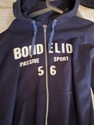 Mörkblå Bondelid hoodie med dragkedja - Mörkblå hoodie från Bondelid med vit text på bröstet och huvan. Modellen har dragkedja, fickor framtill och blå snören i huvan. Perfekt för en avslappnad och sportig look. Materialet är mjukt och bekvämt, med tryck som 'Passive Sport' och 'Offside United'.