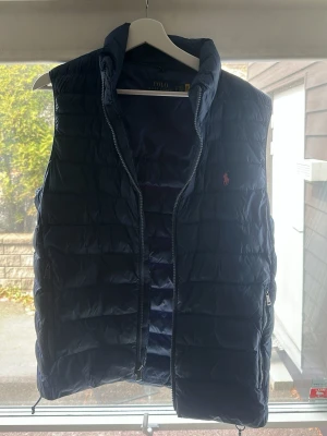 Ralph Lauren väst - Snygg mörkblå dunväst från Polo Ralph Lauren med quiltad design och dragkedja framtill. Västen har hög krage och ikoniska loggan broderad i rött på bröstet. Perfekt för lager-på-lager och håller dig varm under kyliga dagar.