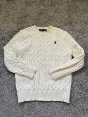Ralph Lauren - Säljer en klassisk vit kabelstickad tröja från Polo Ralph Lauren med rund hals och lång ärm. Tröjan har det ikoniska broderade logotypen på bröstet och är tillverkad i mjukt material som ger en skön känsla. Perfekt för dig som gillar stilrena kläder. 