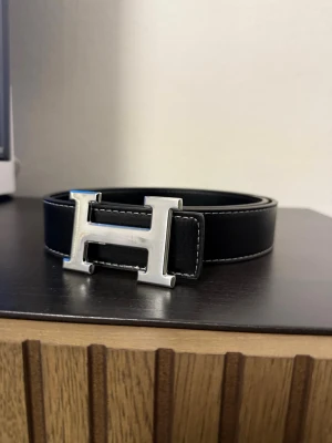 Svart skärp - Stilrent svart skärp. Skärpet har vita kontrastsömmar och är tillverkat i skinn. Perfekt för att lyfta din outfit med en ikonisk detalj.