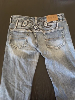Y2k flared bootcut jeans D&G w28-29 - Säljer ett par blå jeans från Dolce & Gabbana med slitna detaljer och broderad D&G-logga på bakfickorna. Jeansen har klassisk femficksdesign och läderpatch med logotyp i midjan. Materialet är denim och färgen är ljusblå med tvättade partier.