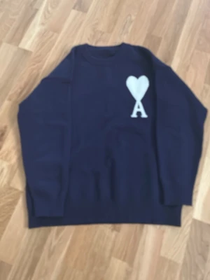 Mörkblå stickad tröja från Ami - Snygg mörkblå stickad tröja från Ami med vit hjärtformad A-logo på bröstet. Tröjan har rund halsringning och ribbade muddar vid ärmslut och nederkant. Materialet är mjukt och skönt, perfekt för att hålla dig varm. Ami paris 