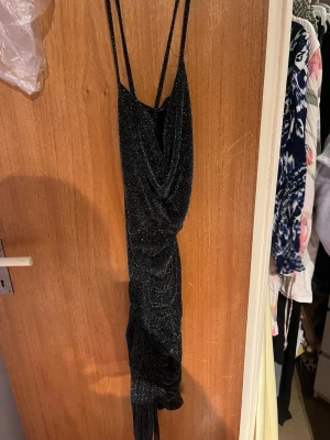 Svart glittrig långklänning - Svart långklänning med tunna axelband och glittrigt material. Klänningen har draperad front och en elegant, slank siluett som framhäver figuren. Perfekt för dig som vill sticka ut med en glamorös look.