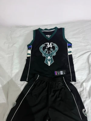 Milwaukee Bucks basketset svart - Säljer ett svart basketset från Milwaukee Bucks med nummer 34 och Antetokounmpo på ryggen. Setet består av ett linne och shorts med gröna och vita detaljer samt Bucks-logga. Perfekt för basketträning eller att representera ditt favoritlag.
