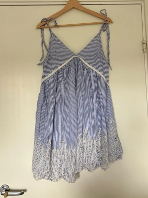 Blåvit randig klänning från ASOS DESIGN - Säljer en kort blåvit klänning från ASOS DESIGN med tunna axelband som knyts. Klänningen har ett randigt mönster och v-ringad front, samt vit spetsdetalj längs nederkanten. 