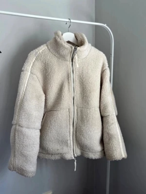 Beige teddyjacka med dragkedja - Mysig teddyjacka i beige med hög krage och hel dragkedja framtill. Jackan har en fluffig och mjuk yta i ett skönt material. Passar perfekt till sommaren/våren.