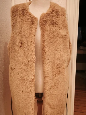Beige fluffig pälsväst med knytskärp Rituals  - Väst i fuskpäls med ett knytband i midjan. Västen har en öppen design framtill och är utan ärmar. Endast provad .