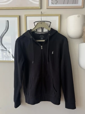 Svart Zip hoodie från Polo Ralph Lauren - Säljer en svart hoodie från Polo Ralph Lauren med dragkedja och huva. Klassisk design med logotypen broderad på bröstet. Hoodien har snören i huvan och praktiska fickor framtill. Materialet är mjukt och bekvämt, perfekt för en avslappnad stil. Pris diskuterbart 