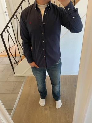 Mörkblå skjorta från Polo Ralph Lauren - Säljer en klassisk mörkblå skjorta från Polo Ralph Lauren med knappar framtill och långärmad design. Skjortan har en liten röd logobrodyr på bröstet och är tillverkad i bomull. Perfekt för dig som gillar stilrena plagg med ikonisk branding.