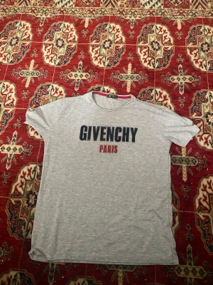 Grå Givenchy Paris t-shirt - Snygg grå t-shirt från Givenchy med stor svart Givenchy-logga och röd Paris-text på bröstet. Klassisk rund halsringning och korta ärmar. Tillverkad i mjuk bomull som känns skön mot huden. Perfekt för dig som gillar stilrena designer med en lyxig touch.