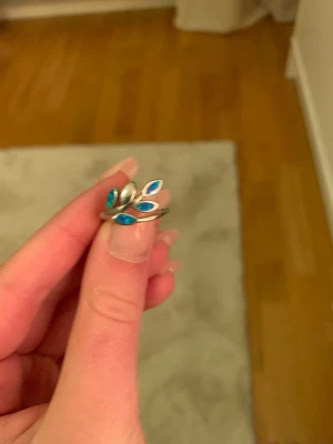 Silverfärgad ring med blå blad - Unik silverfärgad ring med öppet band och dekorativa bladformade detaljer i blått och turkost. Ringen har en modern och stilren design som sticker ut och passar perfekt för dig som gillar färgglada accessoarer. Materialet ser ut att vara metall med emaljerade inslag. Den är rostfri.