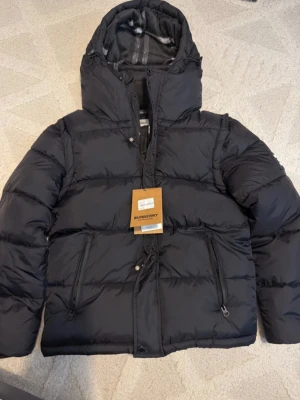 Svart pufferjacka från Burberry - Snygg svart pufferjacka från Burberry med huva och dragkedja framtill. Jackan har två stora fickor med dragkedja och klassiskt rutigt foder i huvan. Perfekt för kalla dagar och riktigt trendig stil.