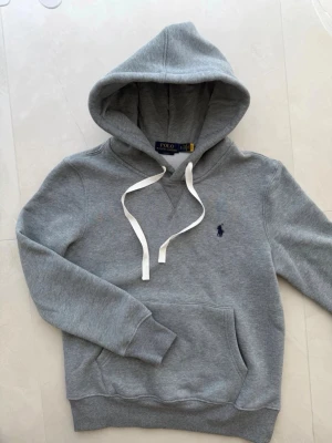 Grå hoodie från Polo Ralph Lauren - Snygg grå hoodie från Polo Ralph Lauren med klassisk huva och vita dragsnören. Tröjan har en känguruficka framtill och den ikoniska mörkblå loggan broderad på bröstet. Tillverkad i mjukt sweatshirtmaterial som är perfekt för chill dagar.