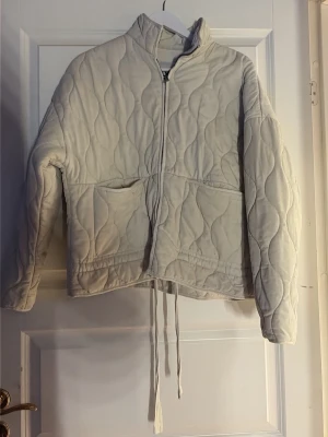 Ljusbeige quiltad jacka med dragsko - Quiltad jacka i ljusbeige med vågigt mönster, dragkedja framtill och två stora fickor. Jackan har hög krage och dragsko i nederkant för justerbar passform. Perfekt för lager-på-lager och stilrena outfits.