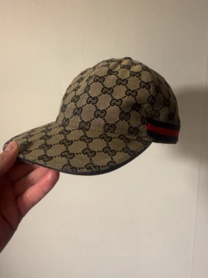 Gucci keps med monogram och röd detalj - Snygg keps från Gucci i beige med klassiskt GG-monogram över hela kepsen. På sidan finns en röd och marinblå rand. Kepsen är tillverkad i Italien och har en böjd skärm. Perfekt accessoar för en trendig streetstyle-look. Kepsen är på perfekt skick och passar size M i huvudform😁