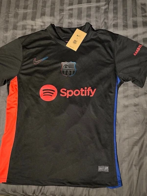 Svart FC Barcelona matchtröja Nike - Ej använd med tagg. Säljer en svart FC Barcelona matchtröja från Nike med Spotify-logga på bröstet. Tröjan har klubbens emblem i svart och detaljer i rött och blått på sidorna. Materialet är lätt och andas, perfekt för fotboll eller träning.