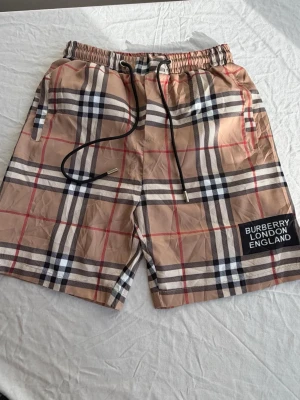 Rutig Burberry shorts med snörning - Snygga shorts från Burberry med klassiskt rutigt mönster i beige, svart, vitt och rött. Shortsen har elastisk midja med svart snörning och en patch med texten 'Burberry London England' på benet. Perfekta för sommaren och riktigt trendiga.