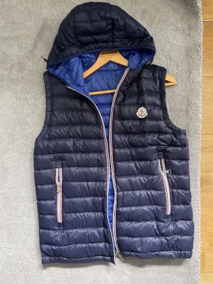 Mörkblå dunväst från Moncler - Snygg mörkblå dunväst från Moncler med avtagbar huva och blått innerfoder. Västen har två fickor med dragkedja och Moncler-logga på bröstet. Klassisk quiltad design och detaljer i rött, vitt och blått vid dragkedjorna. Storlek 3 (M)
