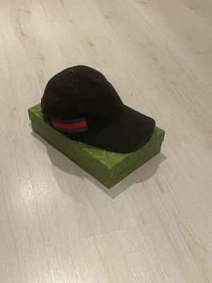 Svart Gucci keps med röd-grön rand - Skickar samma dag innan 20:00 priset kan vi snacka om. Men Snygg svart keps från Gucci med diskret GG-mönster över hela tyget och en klassisk röd-grön rand på sidan. Kepsen har böjd skärm och justerbar rem bak. Perfekt för dig som vill ha en stilren accessoar med lyxig känsla.