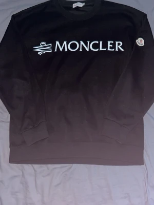 Svart sweatshirt från Moncler - Svart sweatshirt från Moncler med stor vit logga framtill och Moncler-märke på ärmen. Klassisk rund halsringning och ribbade muddar vid ärmslut och nederkant. Perfekt för dig som gillar stilrena och exklusiva plagg.