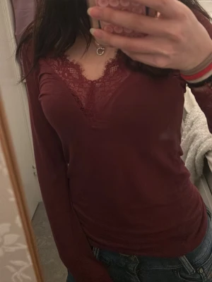 Vinröd långärmad topp med spets - Vinröd långärmad topp med v-ringning och fina spetsdetaljer vid halsen. Sitter tight och har en feminin look som passar perfekt till jeans. Mjuk och stretchig känsla.