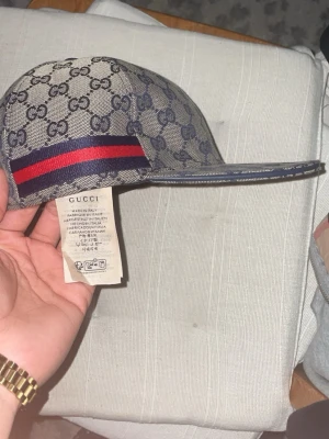 Gucci keps med GG-mönster och röd-blå rand - Snygg keps från Gucci med klassiskt GG-monogram i beige och marinblått. Kepsen har en rak skärm och en ikonisk röd-blå rand på sidan. Tillverkad i canvasmaterial och har märkets logotyp tryckt över hela kepsen. Perfekt för dig som vill sticka ut med en lyxig accessoar.