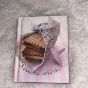 Franska smörkex - Tukan förlag - En söt liten bok med titeln 'Franska smörkex' från Tukan förlag. Omslaget har ett foto på smörkex inslagna i ett blommigt tyg på en rosa- och vitrandig bakgrund. Perfekt för dig som vill baka klassiska franska småkakor.