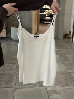 Vit spetsdetaljerad linnet New yourker - Säljer en vit linnetopp från New yourker med tunna axelband och vacker spets längs urringningen. Toppen har en enkel och stilren design som passar perfekt till många olika outfits. Materialet känns mjukt och lätt mot huden.