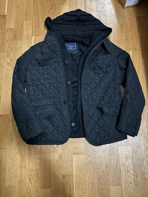 Svart/ Navy blue  quiltad jacka - Roosevelt  - Roosevelt quilted jacket med avtagbar hoodie  En lite schystare jacka med grisch stil – perfekt mix mellan uppklätt och street.  Kan köras utan hood för en clean blazer look eller med hood för mer layered vibe  Quiltat material och mjuk innerfoder , snygg passform och enkel att styla   • Strl L • Färg: svart och navy blue  • Avtagbar hoodie (2 looks i 1) • Väldigt bra skick  • Fickor framtill
