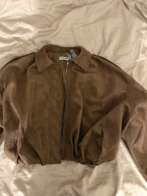 Brun mockajacka från Meet Me There - Snygg brun mockajacka från Meet Me There i modellen Miami Jacket. Jackan har klassisk krage, dragkedja framtill och en loose passform. 