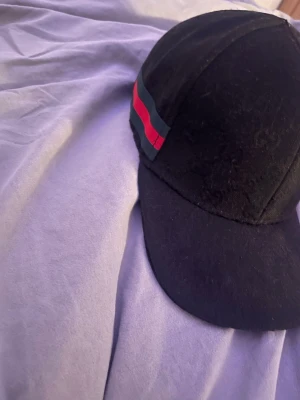 Säljer en svart keps från Gucci med röd-gröna ränder. - Snygg svart keps från Gucci med klassiska röd-gröna ränder på sidan. Kepsen har böjd skärm och justerbart spänne baktill i metall. Materialet är mjukt tyg och kepsen har diskret GG-mönster i tyget för en exklusiv känsla.