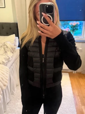 Svart cardiganjacka från Moncler - Snygg svart cardiganjacka från Moncler med quiltad framsida och fyra fickor på varje sida. Jackan har dragkedja framtill, rund halsringning och Moncler-logga på ärmen. Materialmix med vadderad front och stickade ärmar ger en modern look.