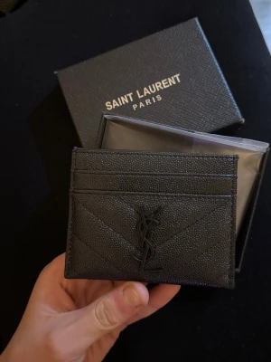Sv definerad korthållare från Saint Laurent - Säljer en svart korthållare från Saint Laurent Paris i präglat läder med ikoniska YSL-loggan framtill. Plånboken har flera kortfack och stilren design med diskreta sömmar. Perfekt för dig som vill ha en lyxig och smidig accessoar.