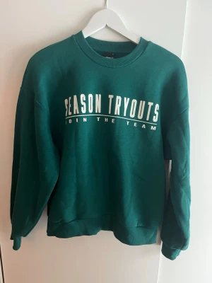 Grön sweatshirt med tryck - Mörkgrön sweatshirt med texten 'SEASON TRYOUTS JOIN THE TEAM' i vitt framtill. Tröjan har rund halsringning, ribbade muddar och är långärmad. Perfekt för en avslappnad och sportig stil. Mjukt material på insidan.