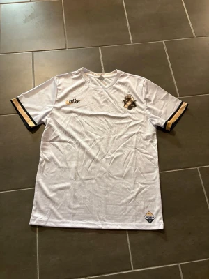 Vit AIK fotbollströja Nike - AIK fotbollströja från Nike i vit med grått grafiskt mönster över hela framsidan. Guld- och svartdetaljer på ärmsluten, AIK-emblem på bröstet och Nike-logga i guld. Materialet är lätt och andas, perfekt för match eller träning.