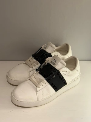 Valentinos skor  - Snygga vita sneakers från Valentino med en bred svart rem över vristen dekorerad med svarta nitar. Skorna har klassisk låg profil, vita snören och en chunky sula. Tillverkade i läder för en exklusiv känsla och cool kontrast mellan vitt och svart.(någon sten är borta men annars bra skick)