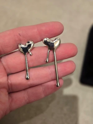 Silvriga hjärtörhängen i droppform - Unika örhängen i blankt silverfärgat metallmaterial med hjärtformad topp och droppformad, asymmetrisk design. Perfekta för dig som gillar statement-smycken med modern twist. 