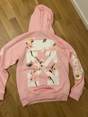 Rosa Off-White hoodie med blommor - Säljer en rosa hoodie från Off-White med stor vit logga och körsbärsblommor på ryggen samt ärmarna. Detaljer som bin och texten 'GLOBAL WARMING?' nedtill. Hoodien har huva med dragsko och magficka. Perfekt statement-plagg för dig som gillar streetwear och unika prints.
