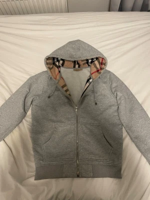 Grå hoodie med Burberry-mönstrad luva - Snygg grå zip-hoodie från Burberry med klassiskt rutigt mönster i luvan. Hoodien har dragkedja framtill, två stora fickor och ribbade muddar. Perfekt för en chill och stilren look.