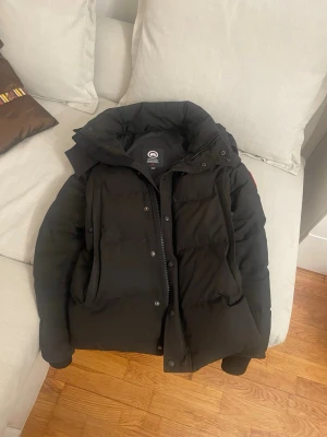 Svart dunjacka från Canada Goose - Säljer en svart dunjacka från Canada Goose med hög krage och stor huva. Jackan har två stora framfickor, dragkedja och tryckknappar. Perfekt för kalla vinterdagar och riktigt varm. Klassisk Canada Goose-logga på ärmen.