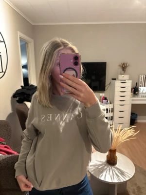 Beige sweatshirt från Cannes - Säljer en beige sweatshirt från Cannes med vit text på bröstet. Tröjan har rund hals, ribbade muddar vid ärmslut och nederkant samt en avslappnad passform. Perfekt för en chill look och enkel att matcha med jeans eller leggings.
